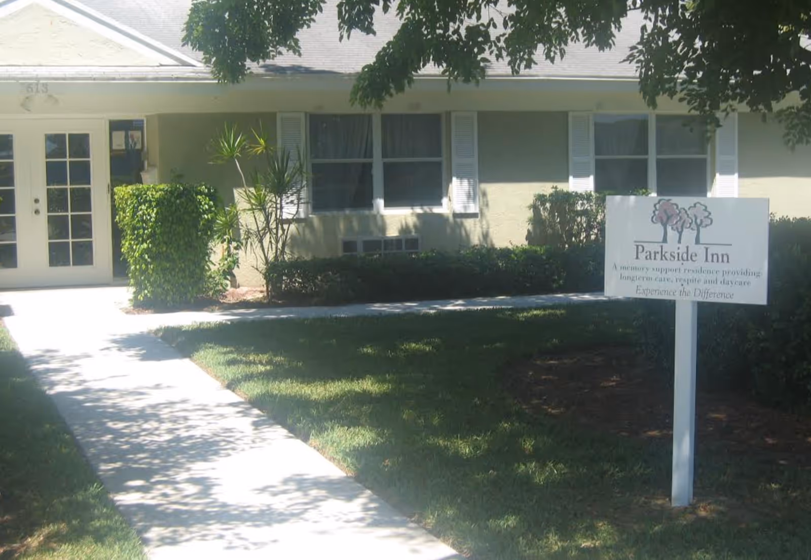 Exterior view of a single-story building with a walkway leading to a double glass door entrance. There are bushes and trees casting shadows on the grass lawn. A sign in front reads 'Parkside Inn, A memory support residence providing longterm care, respite and daycare. Experience the Difference.'