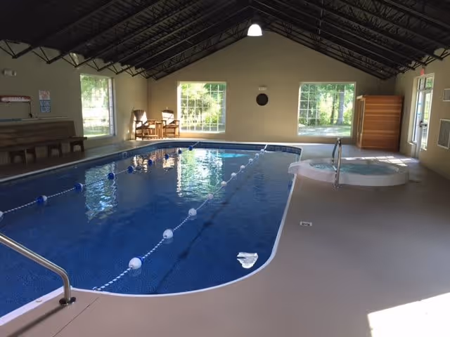 Indoor swimming pool with lane dividers and a small round hot tub adjacent to it inside a room with large windows and a high ceiling with exposed beams. There are chairs and benches along the walls, and sunlight is streaming through the windows.