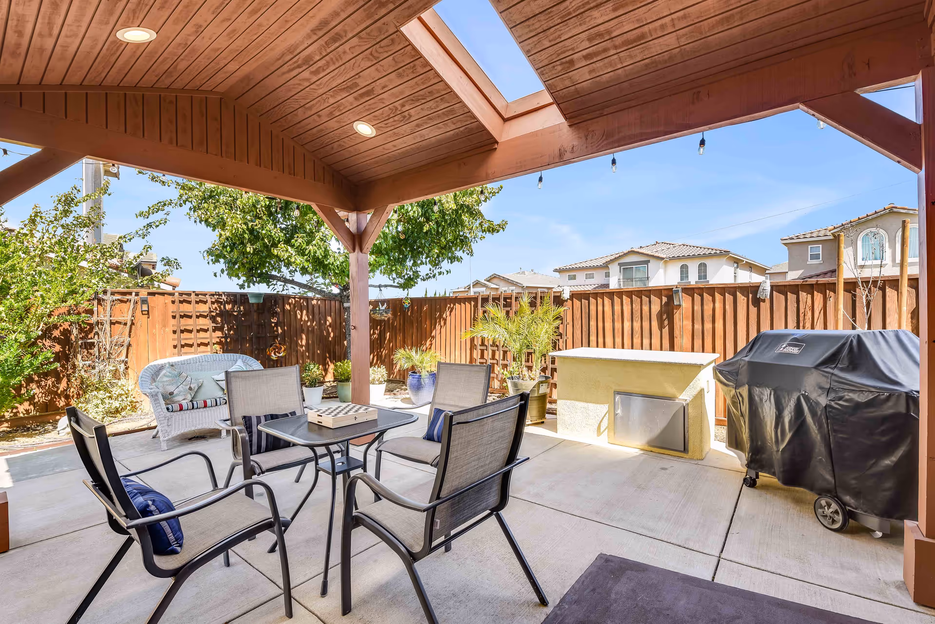 Covered outdoor patio area with a wooden ceiling and skylight, featuring a table with four chairs, a white wicker loveseat with cushions, potted plants, a built-in outdoor fireplace, and a covered barbecue grill. The patio is enclosed by a wooden fence with neighboring houses visible in the background under a clear blue sky.
