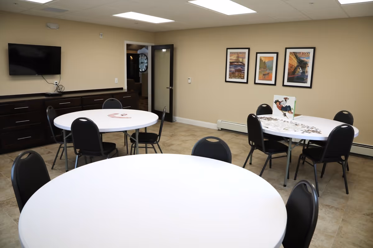 Interior room with three round white tables surrounded by black chairs. One table has a puzzle in progress with a puzzle box on it. The room has beige walls, a tiled floor, a flat screen TV mounted on the wall, dark wood cabinets, and three framed pictures hanging on the wall.