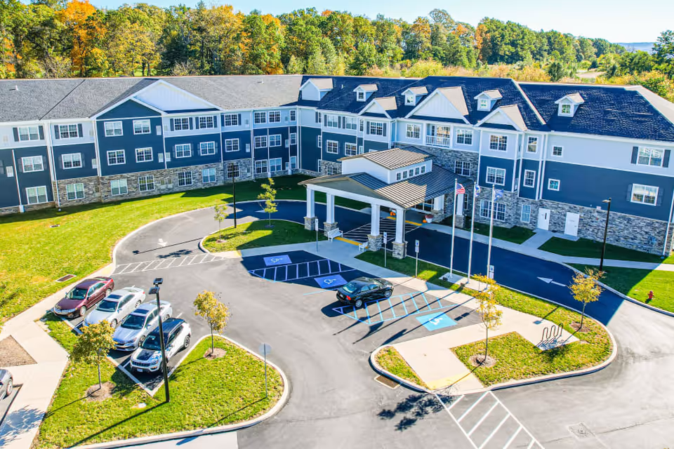 Aerial view of a three-story senior living building with a covered main entrance, parking lot with handicapped spaces, and surrounding trees.