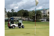 A golf cart parked on a green golf course near a hole with a flag. In the background, there are trees and a large multi-story building under a partly cloudy sky.