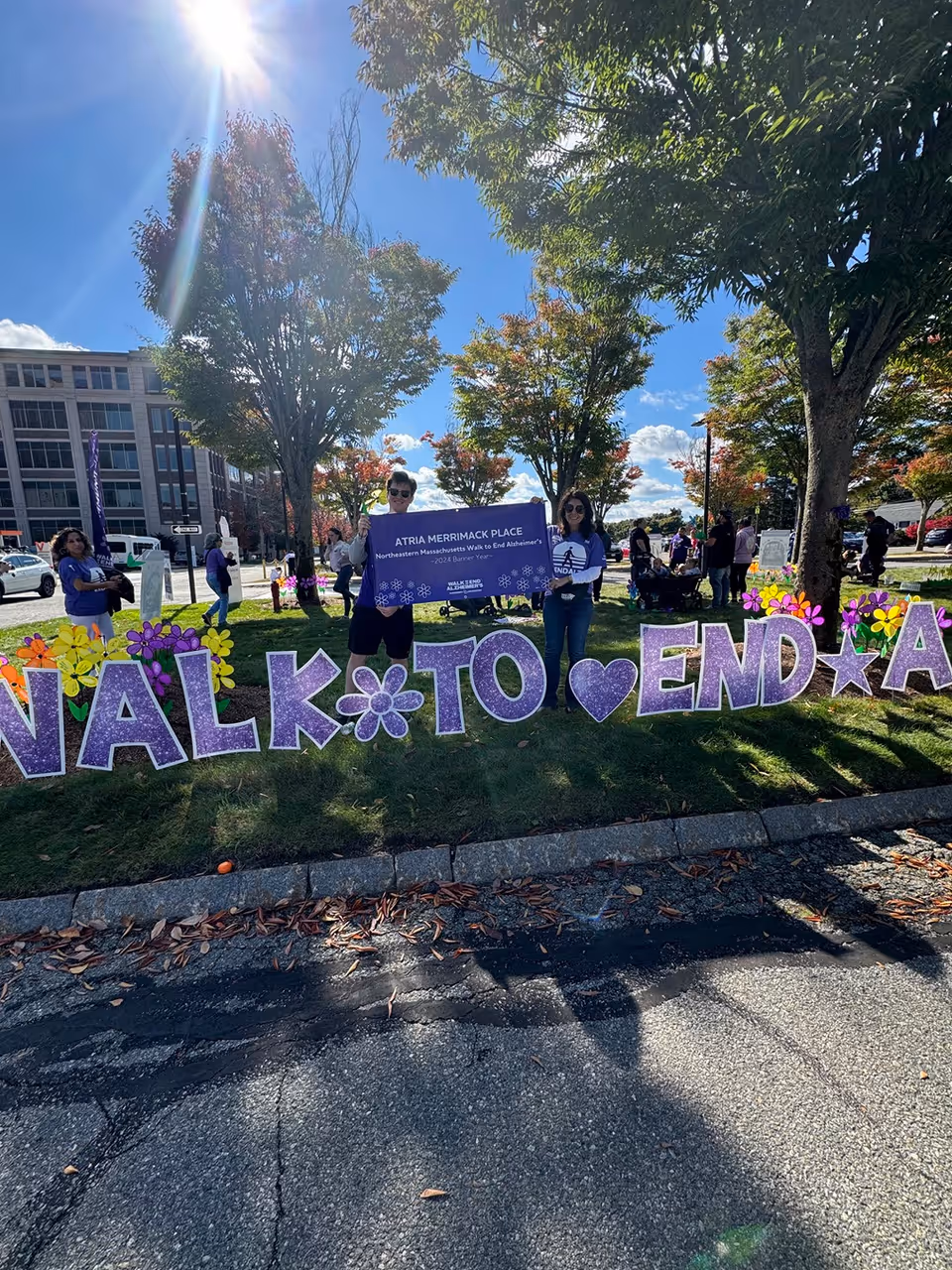 Two people standing outdoors on a sunny day holding a purple banner that reads 'Atria Merrimack Place Northeastern Massachusetts Walk to End Alzheimer's 2024 Banner Team'. They are standing behind large purple letters and symbols on the grass that spell out 'WALK TO END A'. Trees and other people are visible in the background.