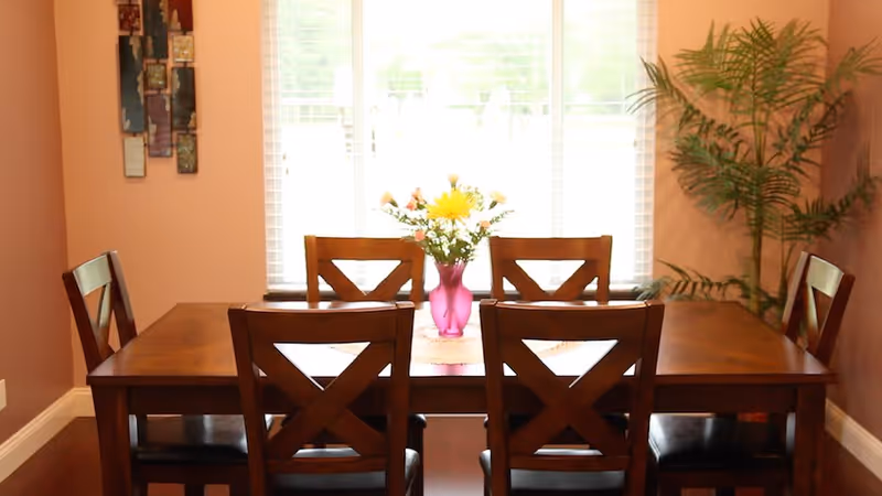 A wooden dining table with six matching chairs arranged around it. A pink vase with yellow and white flowers is placed in the center of the table. Behind the table is a large window with white blinds, and to the right is a tall green potted plant. The walls are painted a warm beige color, and there is a decorative wall hanging on the left side.