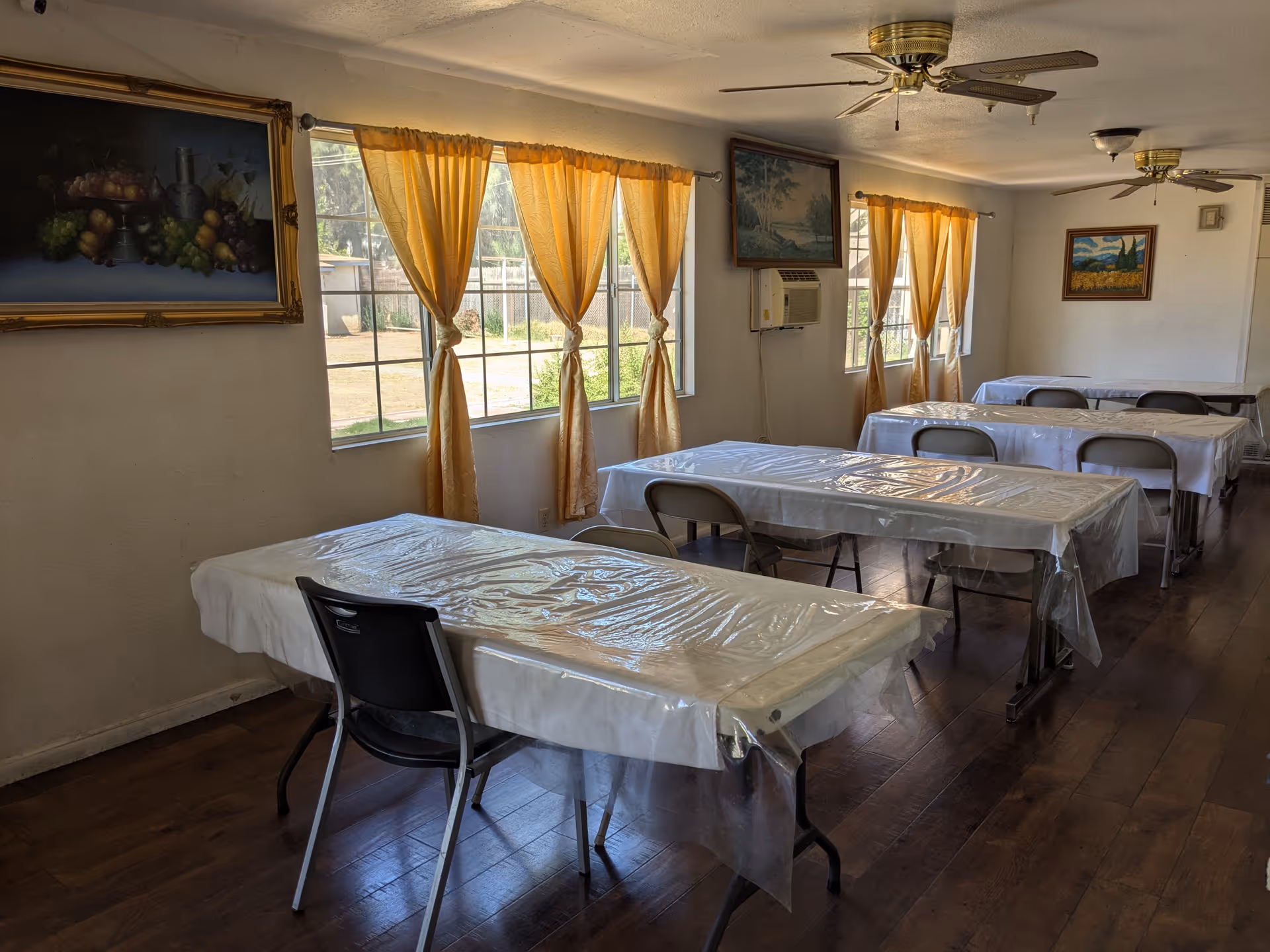 Long dining room with several rectangular tables covered in plastic, folding chairs, yellow-curtained windows, ceiling fans, and framed paintings on the walls.