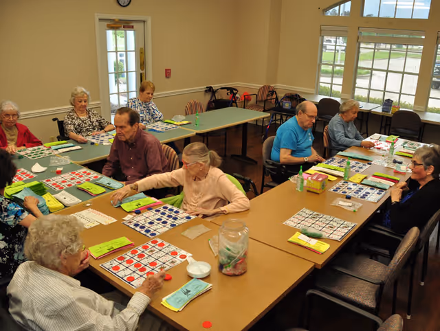 A group of elderly people sitting around tables in a well-lit room playing bingo. The room has large windows, beige walls, and several chairs. Bingo cards and markers are spread out on the tables.
