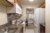 A narrow kitchen with wooden cabinets, a white microwave above a stove with four burners, a stainless steel sink, and a white refrigerator. The kitchen opens into a living room area with a ceiling fan and a window with curtains.