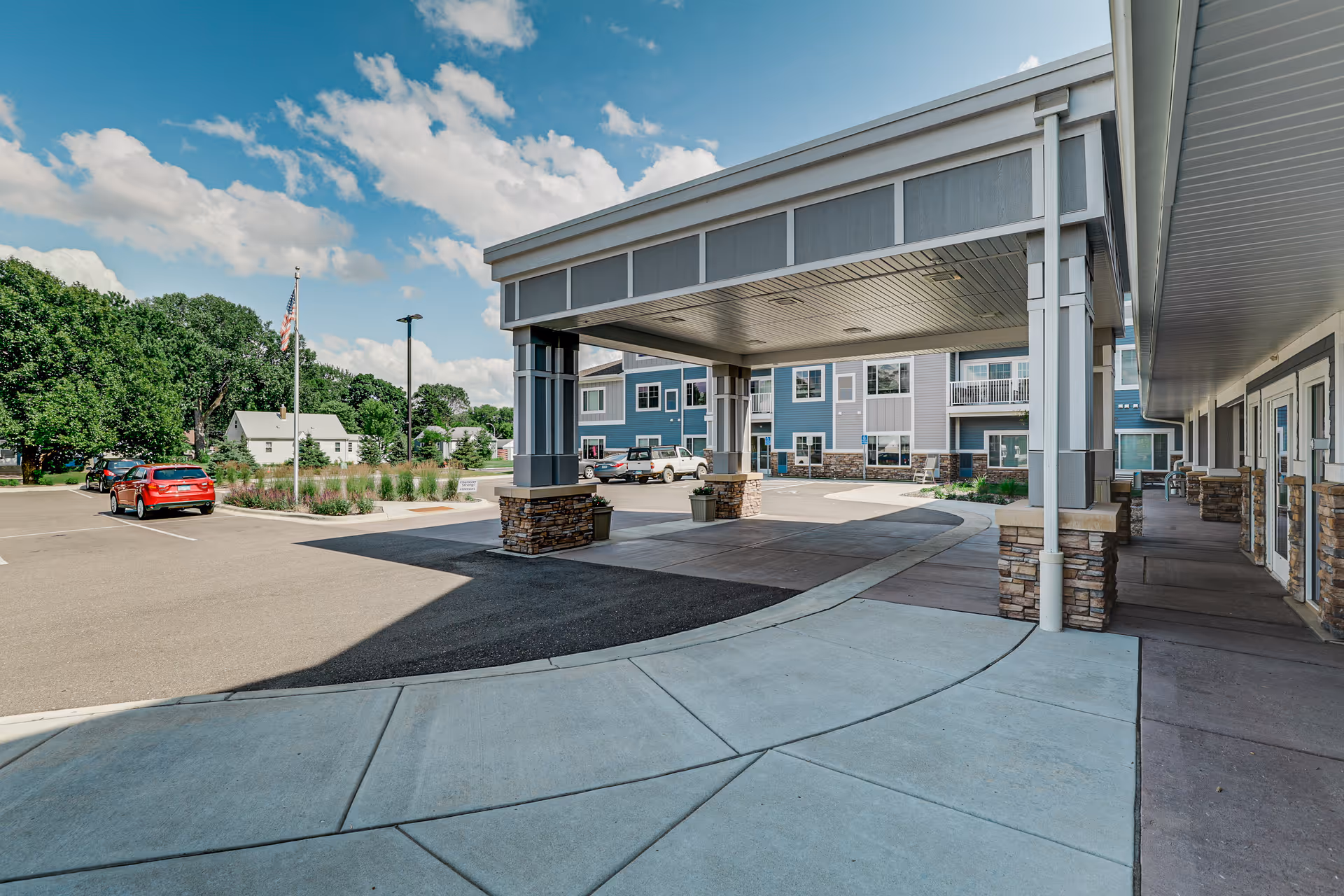 Covered entrance and porte-cochere of a senior living building with parked cars, sidewalk, and an American flag.