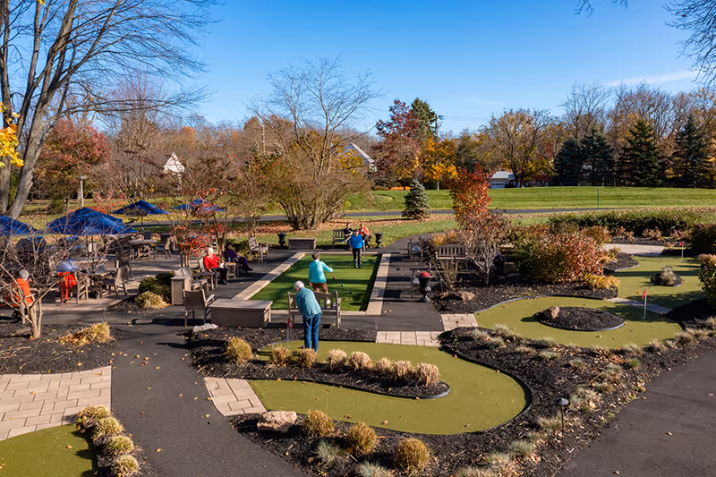 Outdoor recreational area at Gwynedd Estates featuring a mini-golf course and bocce ball court surrounded by trees with autumn foliage. Several seniors are playing and sitting under blue umbrellas at tables and chairs. The sky is clear and blue.