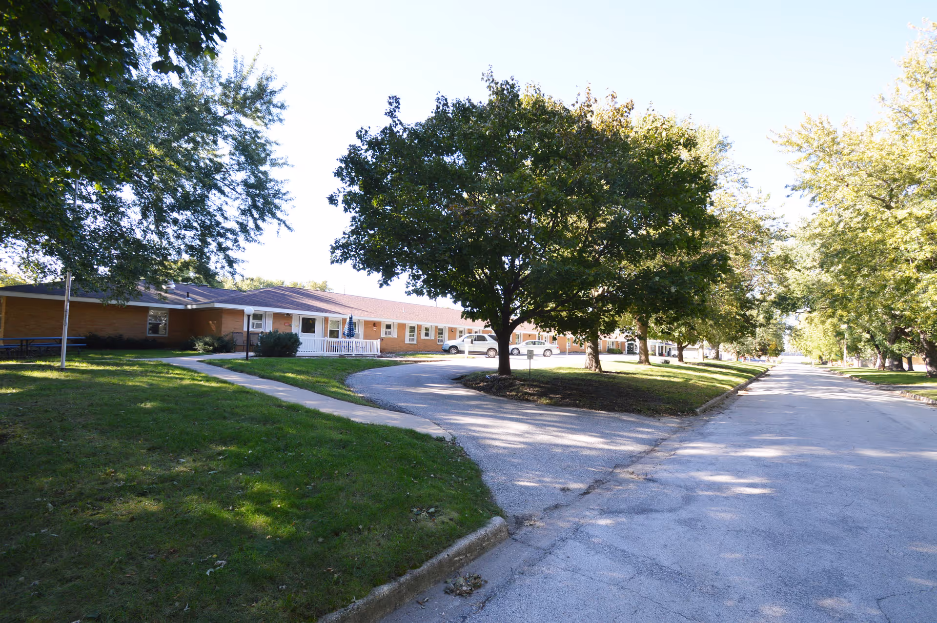Exterior view of a single-story brick building with a sloped roof, surrounded by green grass and large trees. A paved driveway curves in front of the building, and a road runs alongside it. Several parked cars are visible near the building.