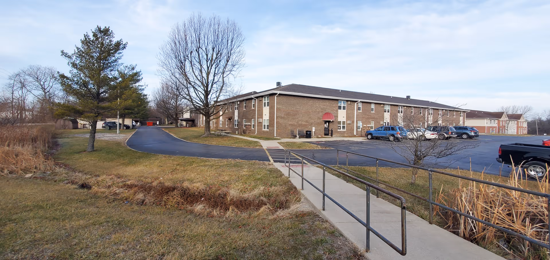 Exterior view of Cougill Senior Apartments showing a two-story brick building with multiple windows, a red awning above the entrance, a parking lot with several cars, a paved driveway, leafless trees, and a concrete ramp with metal railings leading up to the building. The sky is partly cloudy.