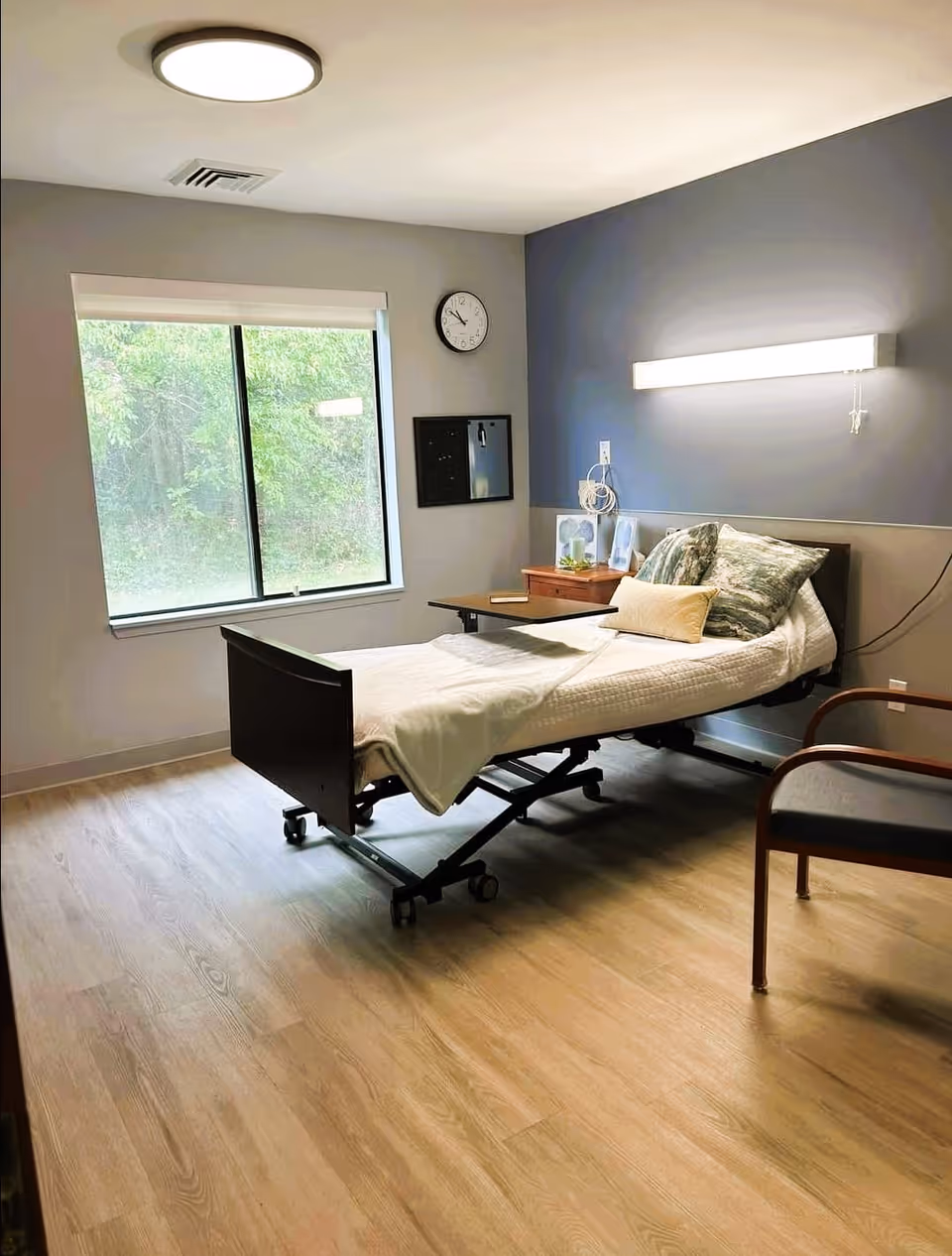 A clean nursing facility bedroom with an adjustable hospital bed, bedside table, chair, large window, and wall-mounted light.