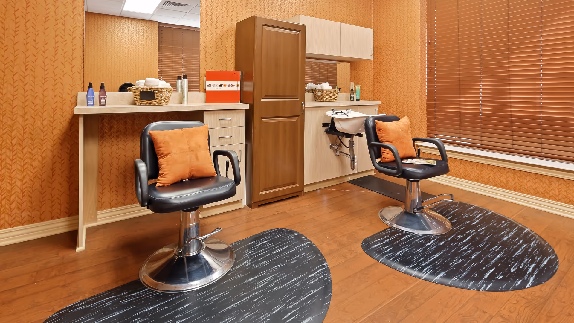 Interior of a salon room with two black salon chairs each with an orange cushion, a wooden floor, orange patterned wallpaper, a large mirror, a sink for washing hair, and a window with closed brown blinds.
