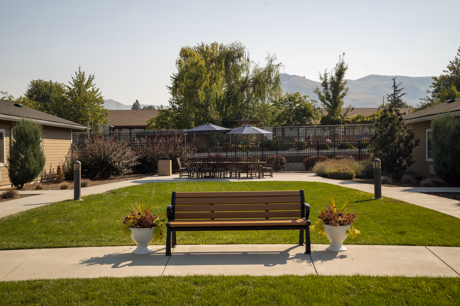 Outdoor courtyard area with a wooden bench flanked by two white planters with flowers, surrounded by green grass and concrete walkways. In the background, there are patio tables with umbrellas, trees, shrubs, and buildings under a clear sky with mountains visible in the distance.