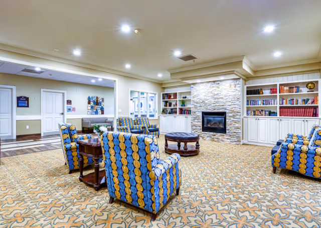 Well-lit senior living common room with patterned armchairs arranged around a central table, built-in bookshelves, and a stone fireplace.