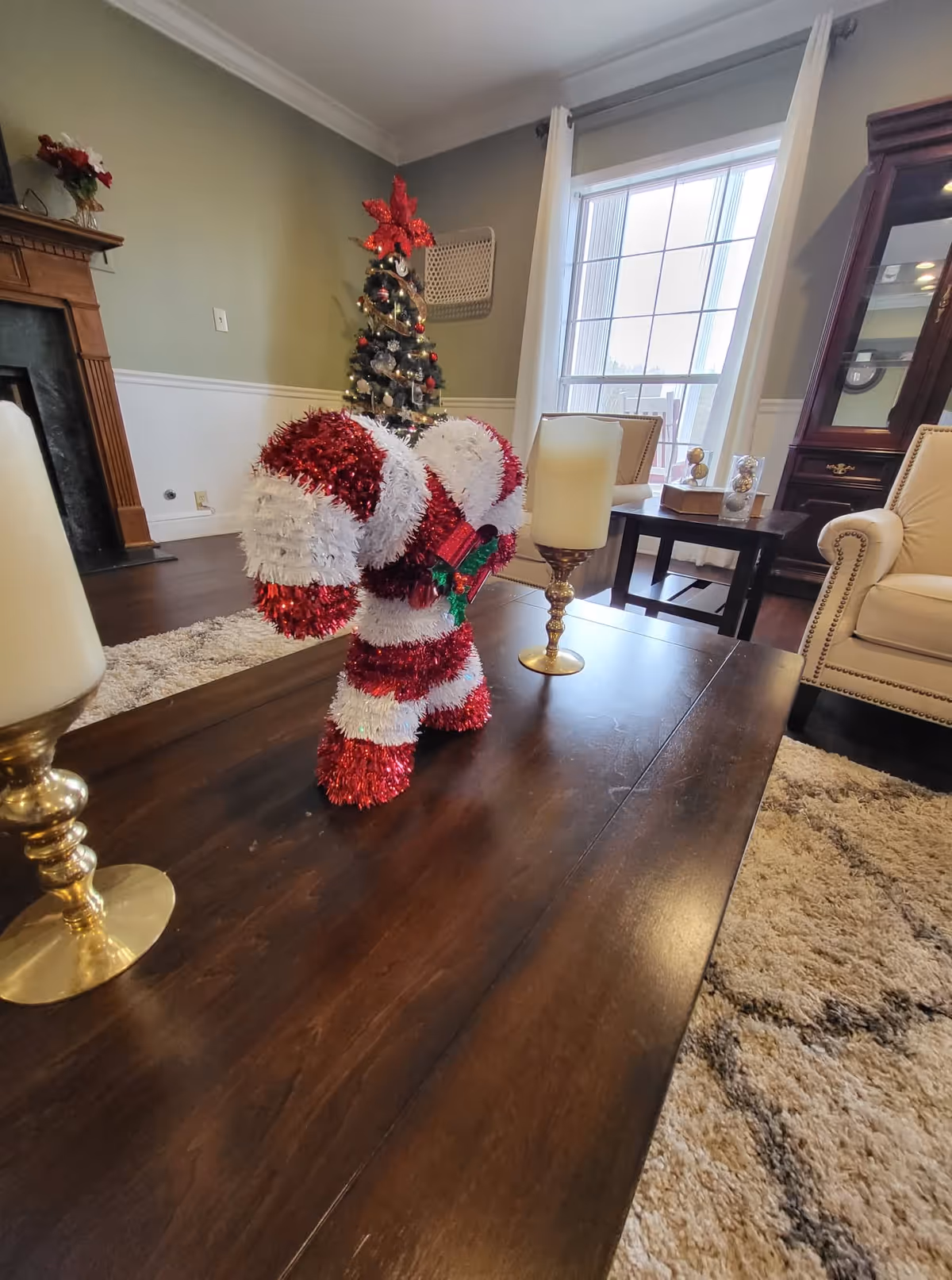 A cozy living room decorated for Christmas with a small Christmas tree in the corner and a red and white striped candy cane decoration on a dark wooden coffee table. The room features beige armchairs, a large window with white curtains, a fireplace, and a beige patterned rug.