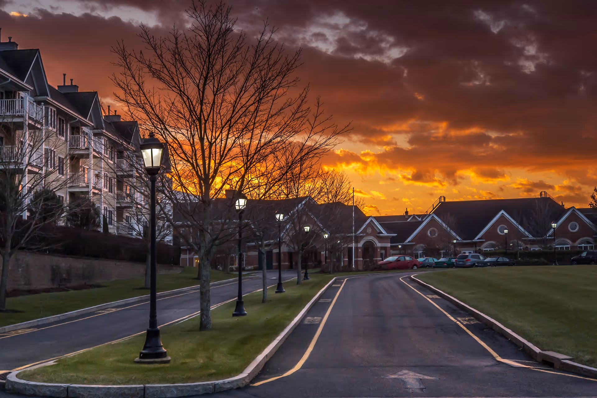 A paved driveway lined with street lamps and leafless trees leads to a large residential building complex under a dramatic sunset sky with orange and purple clouds.