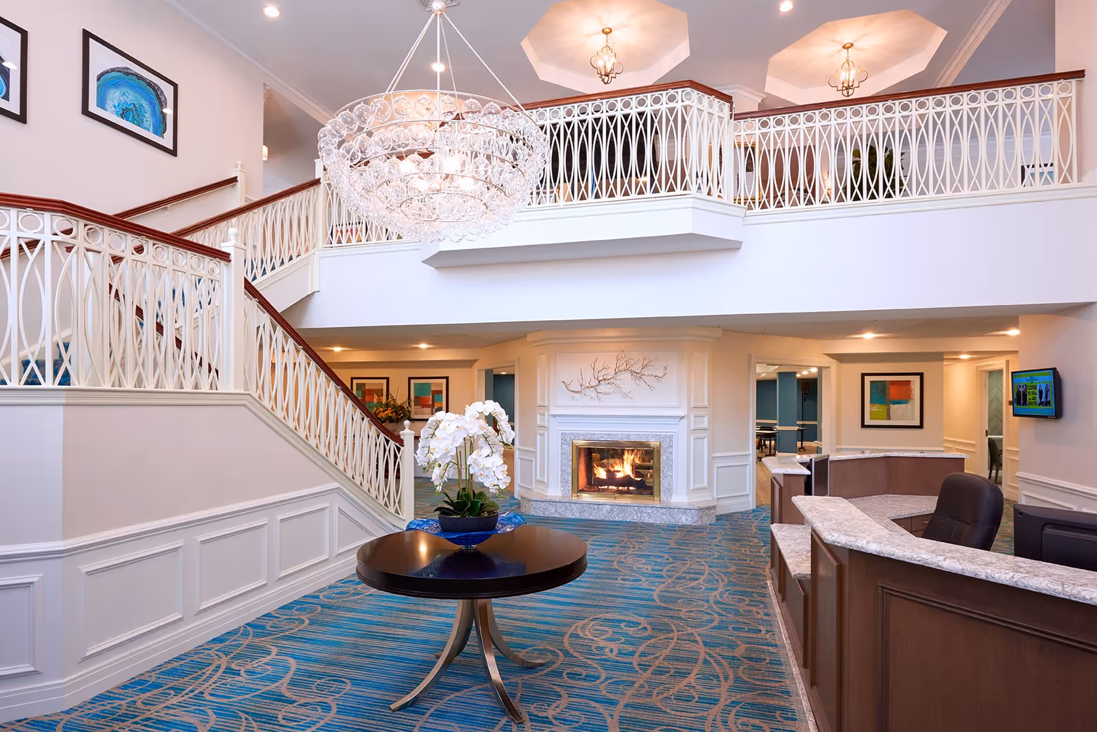 Interior view of a senior living facility lobby featuring a large chandelier, a round table with a white orchid centerpiece, a staircase with white decorative railings, a fireplace with a decorative branch above it, and a reception desk with a computer monitor. The carpet is blue with a swirling pattern, and framed artwork is visible on the walls.