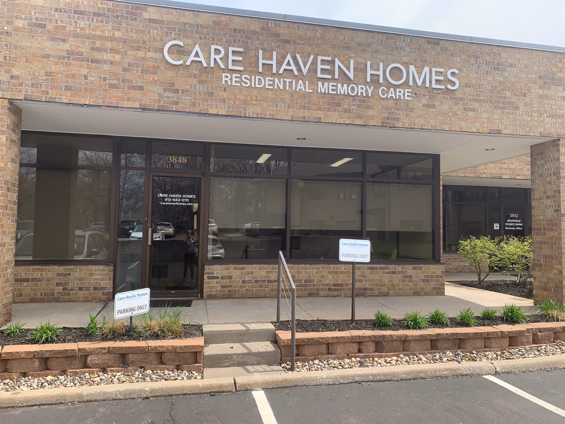 Exterior view of Care Haven Homes building with a brick facade and large windows. The sign above the entrance reads 'CARE HAVEN HOMES RESIDENTIAL MEMORY CARE'. There are two small signs in front of the building indicating parking only for Care Haven Homes. A small set of stairs with a handrail leads to the entrance door.