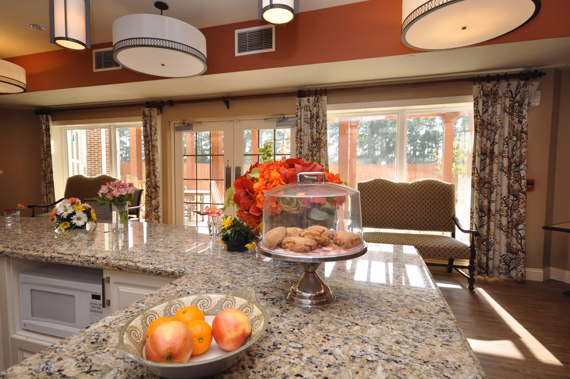 Interior view of a bright room with a granite countertop island featuring a bowl of apples and oranges, a glass-covered plate of cookies, and several flower arrangements. In the background, there are large windows and glass doors with patterned curtains, a cushioned bench, and wooden flooring.