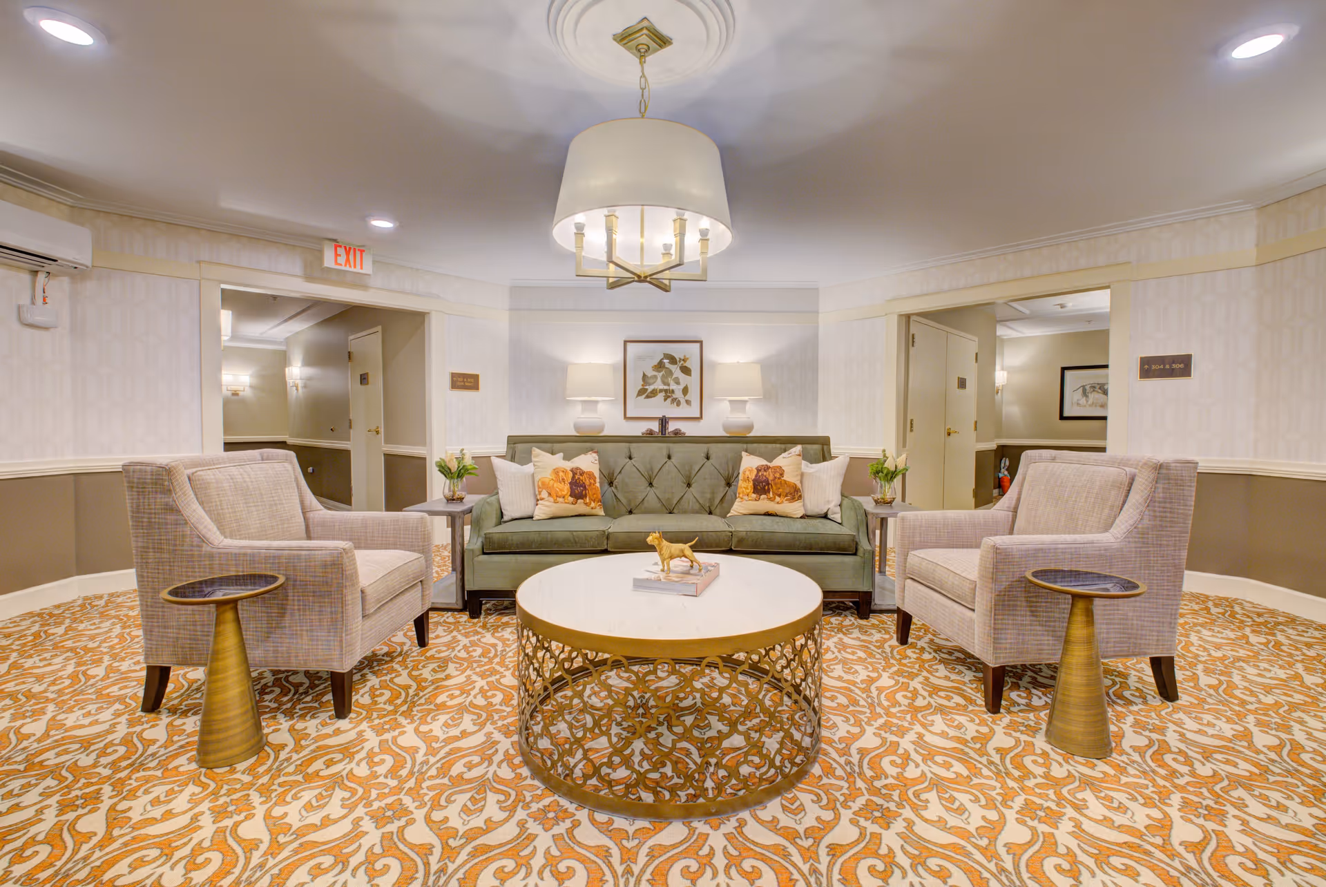 A cozy sitting area in a senior living facility with a green tufted sofa adorned with floral pillows, two beige armchairs, a round coffee table with decorative items, and a patterned carpet. The walls are light-colored with subtle wallpaper, and there are two table lamps on a console behind the sofa. A modern chandelier hangs from the ceiling, and there are doorways leading to other rooms.
