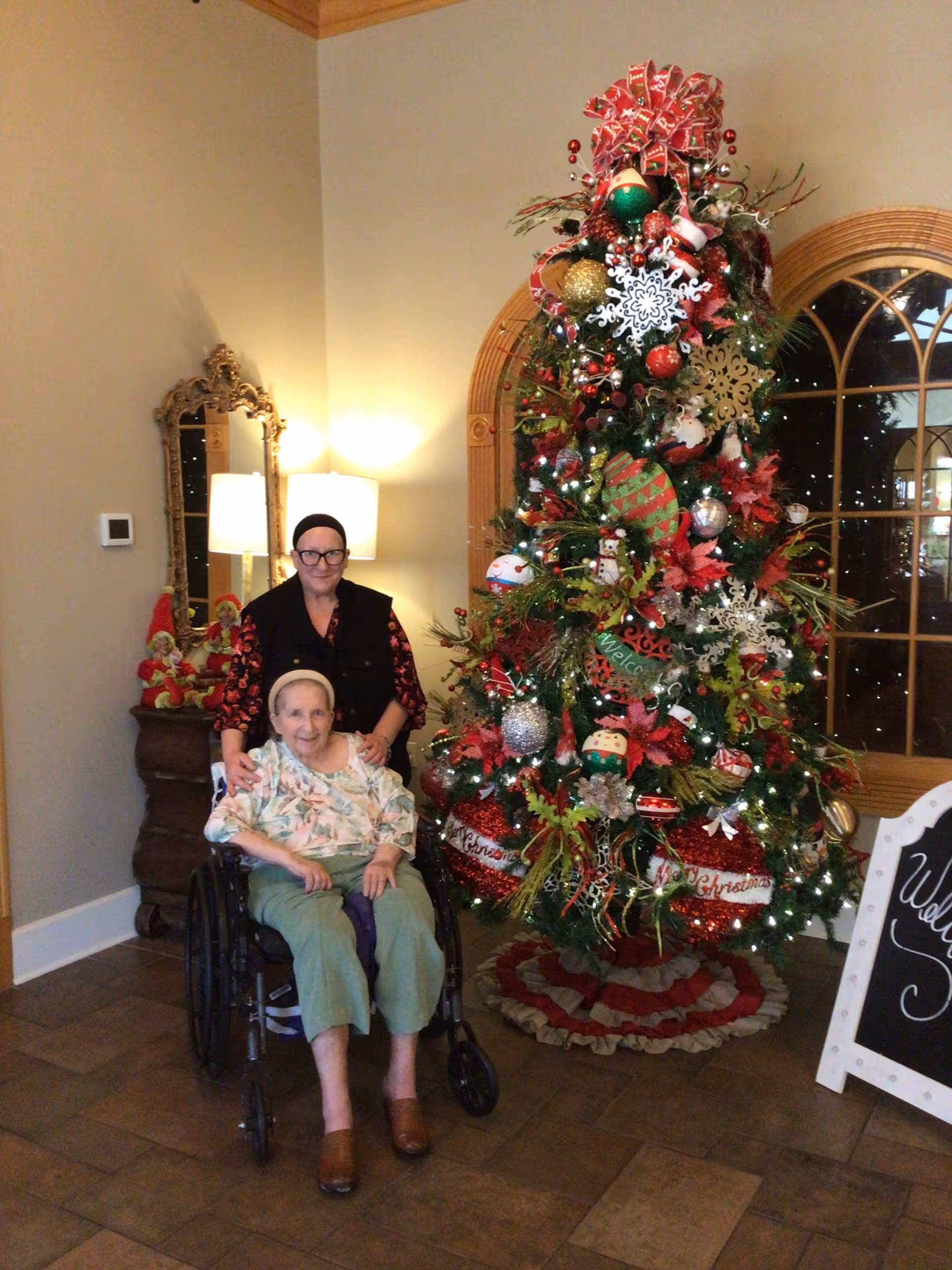 An elderly woman sitting in a wheelchair with a younger woman standing behind her, both smiling, next to a decorated Christmas tree inside a facility. The tree is adorned with ornaments, ribbons, and lights. There is a mirror and a lamp on a wooden dresser in the background, and a chalkboard sign partially visible on the right side.