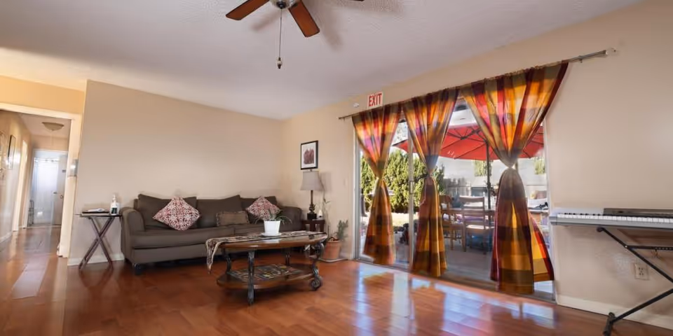 Living room with a sofa, coffee table, ceiling fan and sliding glass doors with colorful curtains opening to a patio.