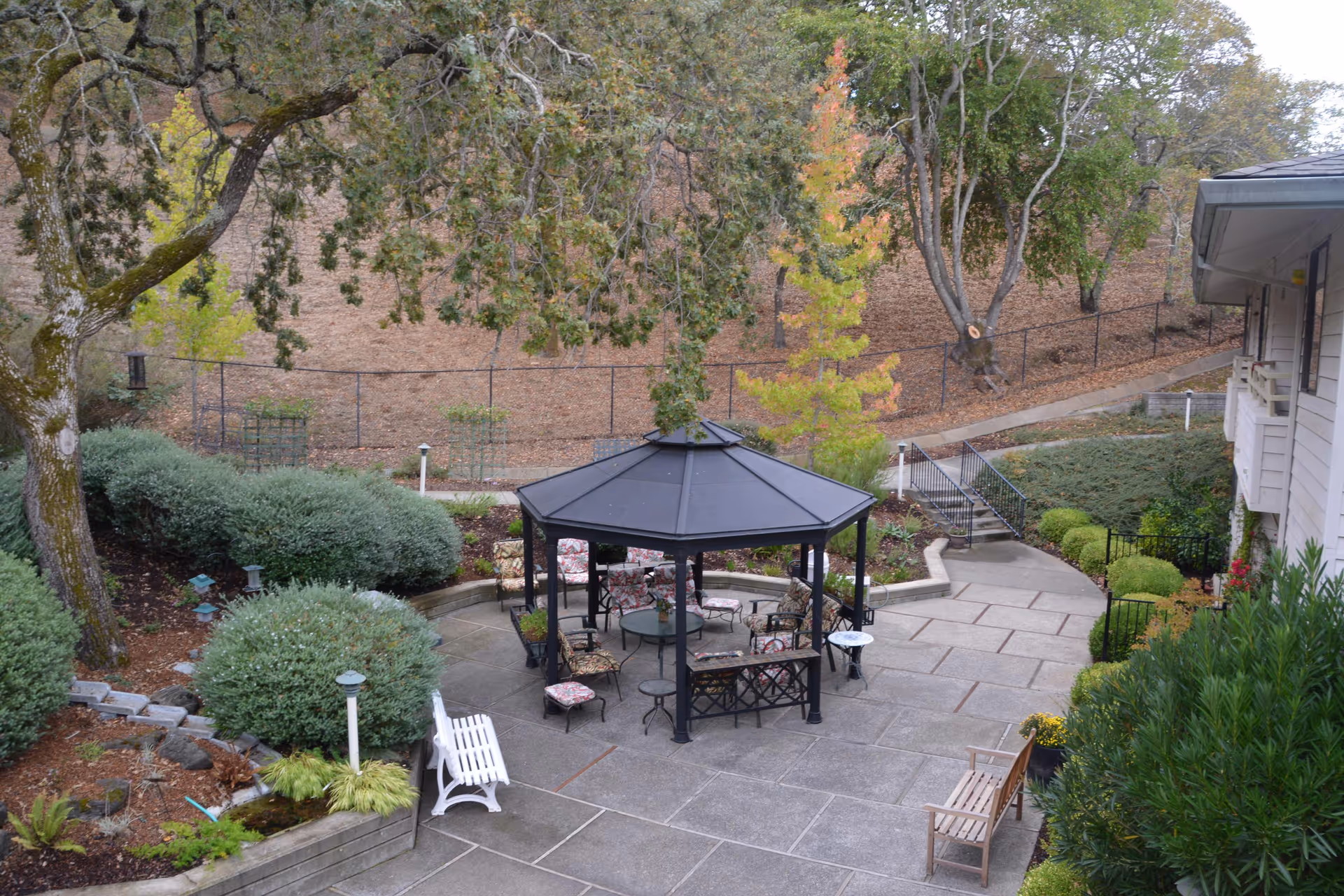 Outdoor patio area at Deer Park Retirement Community featuring a black gazebo with cushioned chairs and a round table underneath. Surrounding the patio are benches, landscaped bushes, trees, and a pathway leading up a small hill with a fence in the background.