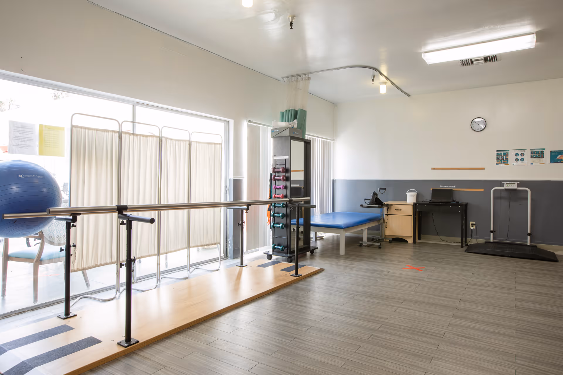 A physical therapy room with parallel bars for walking exercises, a large blue exercise ball, a folding privacy screen, a treatment table, a rack with small dumbbells, a desk with a computer, and a treadmill. The room has large windows letting in natural light and a clock on the wall.