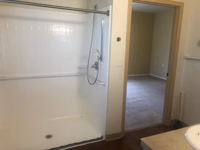 A clean, white walk-in shower with grab bars and a handheld showerhead. The shower has a tiled wall and a metal curtain rod. Next to the shower is a doorway leading to a carpeted room with beige walls. Part of a bathroom sink and countertop is visible on the right side of the image.