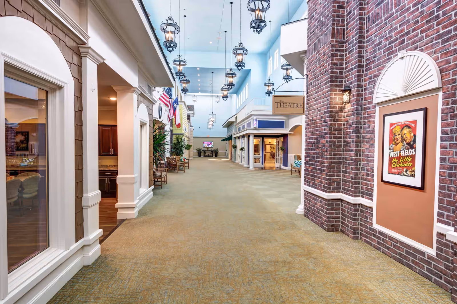 A spacious indoor hallway designed to resemble a small town street with storefronts, benches, and hanging lantern-style lights. The walls feature various architectural styles including brick and siding, with flags displayed on the left side. A sign reading 'The Theatre' hangs on the right side above a doorway, and a vintage movie poster is mounted on a brick wall. The ceiling is high with multiple windows allowing natural light.