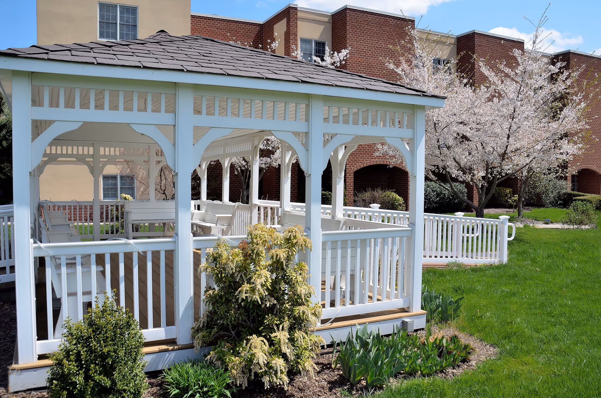 A white wooden gazebo with a shingled roof situated in a green garden area with blooming trees and shrubs. In the background, there is a brick building with windows and archways.