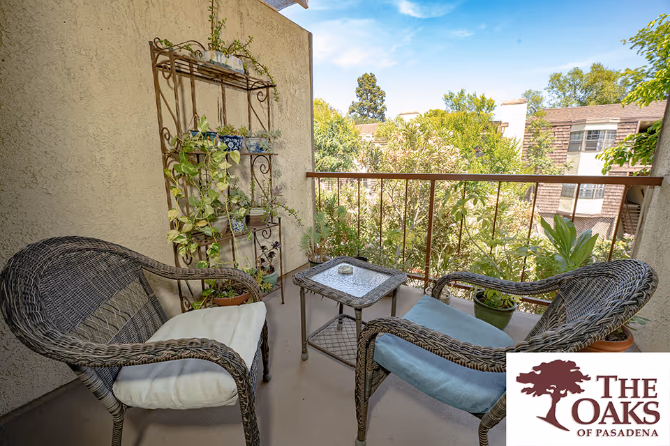A small outdoor balcony area with two wicker chairs with cushions and a small glass-top table between them. There is a metal plant stand with various potted plants against the wall, and more potted plants on the floor. The balcony overlooks green trees and nearby buildings under a clear blue sky.