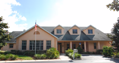 Front exterior of a peach-colored assisted living building with a gabled roof, three dormer windows, a flagpole, and landscaped entrance.