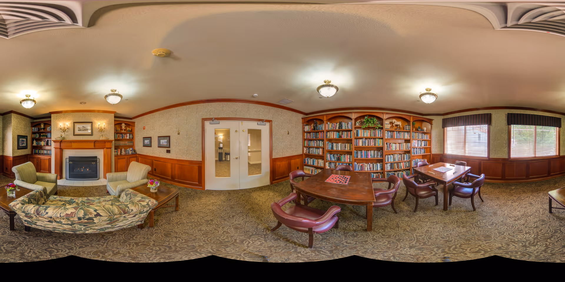 A cozy senior living facility common room featuring a fireplace with two armchairs and a floral-patterned sofa arranged around a coffee table. To the right, there are bookshelves filled with books and two tables with chairs near windows with blinds, allowing natural light to enter. The room has warm wood paneling and carpeted floors.