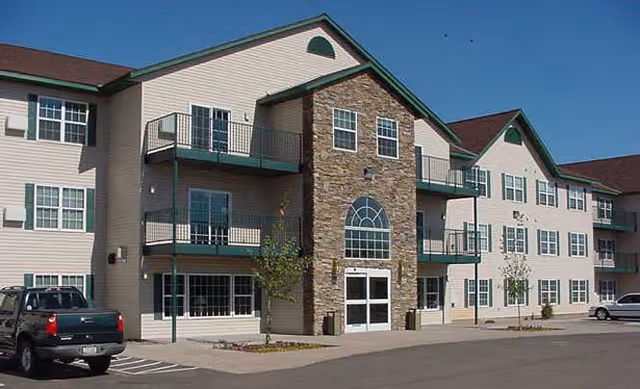 Exterior view of a three-story senior living facility building with beige siding and stone accents around the main entrance. The building has multiple windows and balconies, with a parking lot in front containing a few vehicles. The sky is clear and blue.