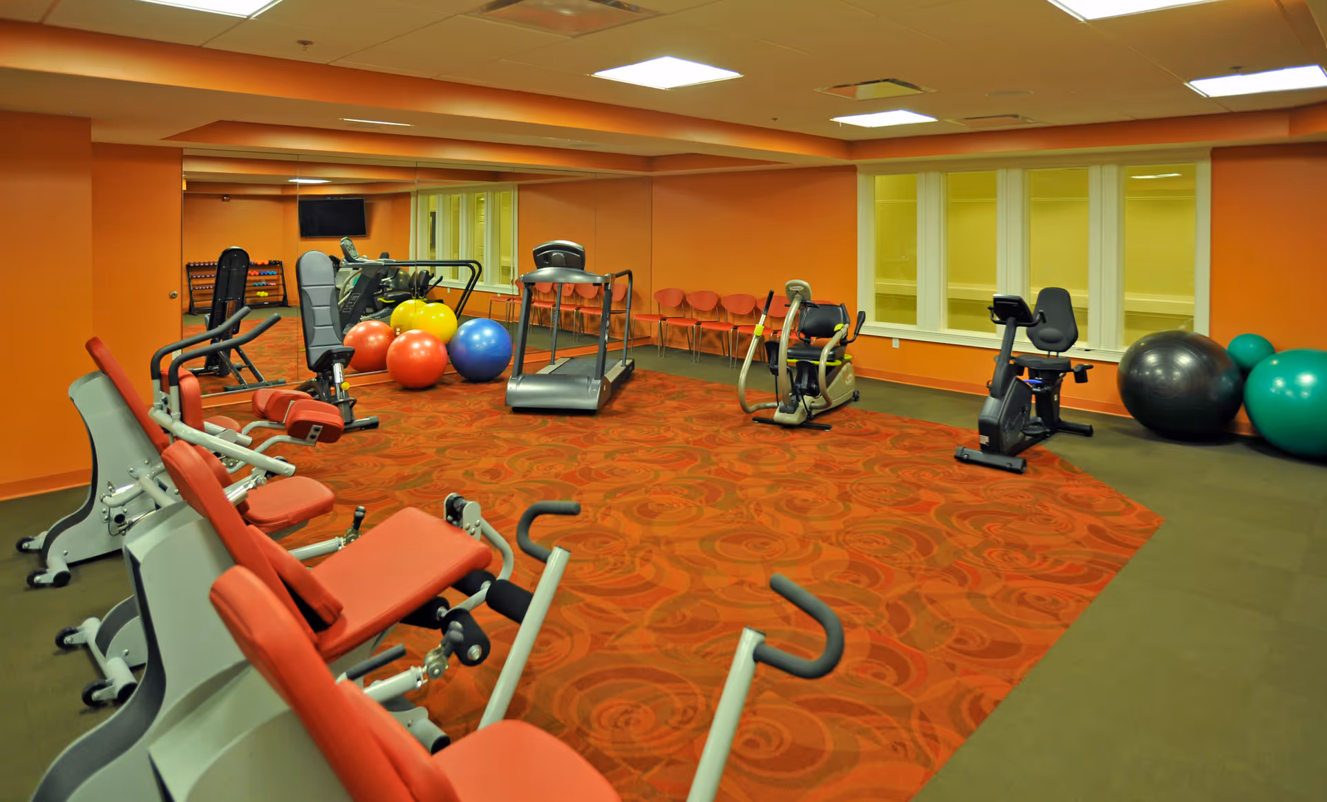 A fitness room with various exercise equipment including stationary bikes, a treadmill, and weight machines. There are colorful exercise balls on the floor and a row of orange chairs along the back wall. The walls are painted orange and there is a large mirror on one side of the room.