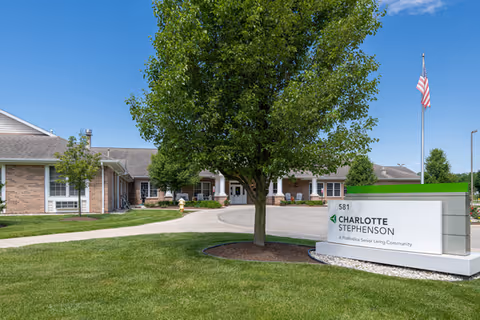 Exterior view of Charlotte Stephenson Manor, a senior living community building with a brick facade, surrounded by green grass and trees under a clear blue sky. A sign in front displays the name and address, with an American flag on a pole nearby.