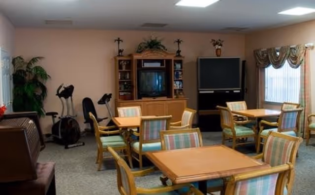 A common area in a senior living facility with multiple wooden tables and chairs with pastel-colored cushions. There is a wooden entertainment center with a TV and shelves, another large TV on a stand, exercise equipment in the corner, and a window with curtains letting in natural light.