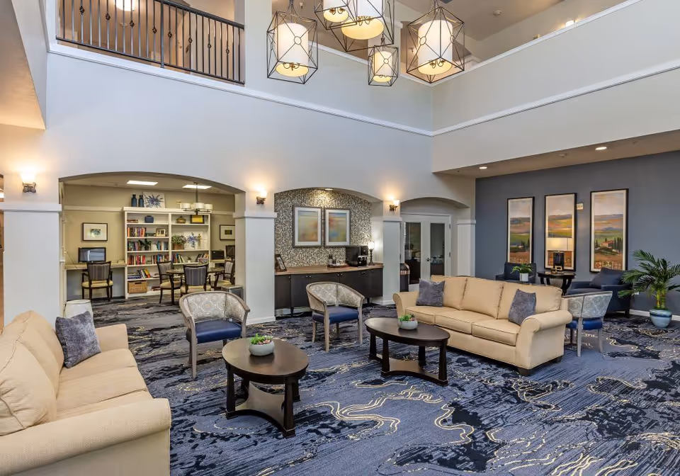 A spacious senior living facility common area with beige sofas, patterned armchairs, and dark wooden coffee tables on a blue patterned carpet. The room features high ceilings with modern hanging light fixtures, a small kitchenette or coffee station with framed artwork above, and a separate room with bookshelves and desks visible through an open archway.