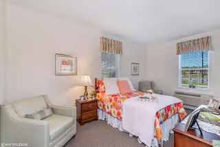 A bright and cozy bedroom with a single bed covered in a floral bedspread and white blanket. The room features two windows with striped valances, a wooden nightstand with a lamp, a comfortable armchair, and framed artwork on the walls. There is also a wooden dresser with decorative items and a heating/cooling unit beneath one window.