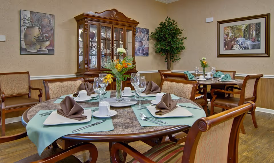 A furnished dining room with round tables set for a meal, folded napkins, glassware, floral centerpieces, chairs, and a china cabinet.