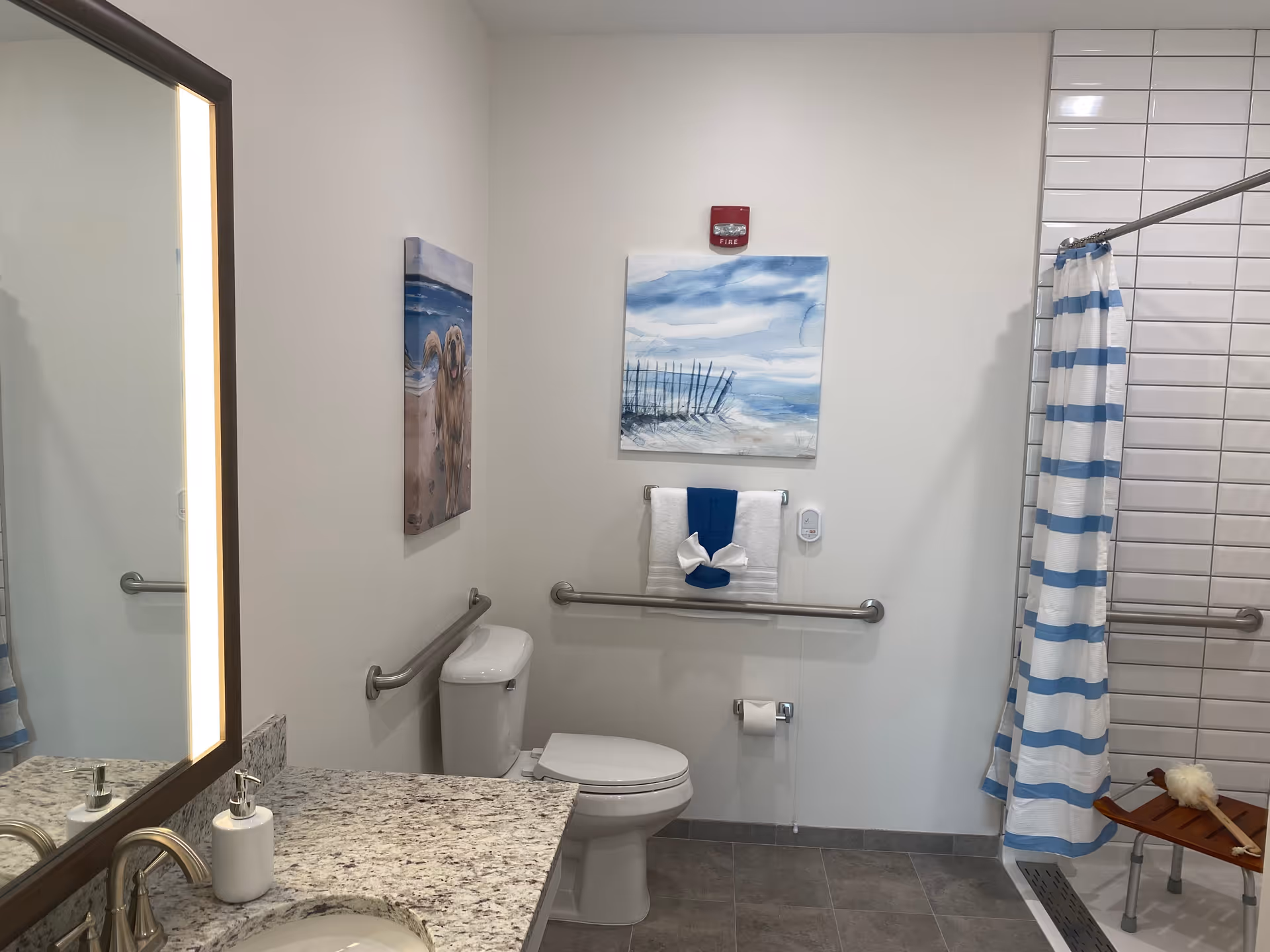 Accessible bathroom with a toilet, granite sink countertop, grab bars, and a shower with a blue-striped curtain and coastal wall art.