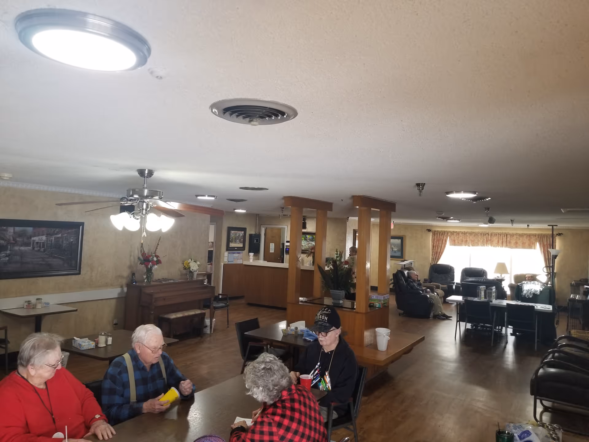 A common area in an assisted living facility with elderly residents sitting around a table engaged in activities. The room has wooden flooring, a ceiling fan with lights, a piano against the wall, and comfortable seating near a window with curtains. There are plants and framed pictures decorating the space.