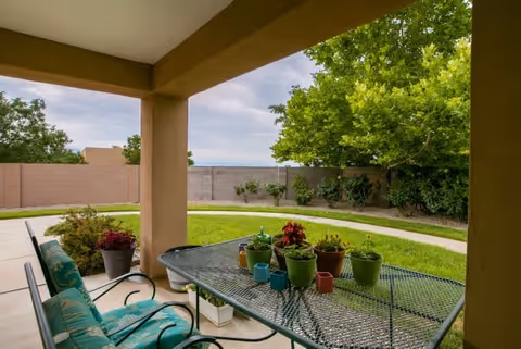 Covered outdoor patio area with a metal table and chairs. The table has several small potted plants on it. Beyond the patio is a grassy yard with a curved concrete pathway, some shrubs, and a tall tree near a beige brick wall under a cloudy sky.