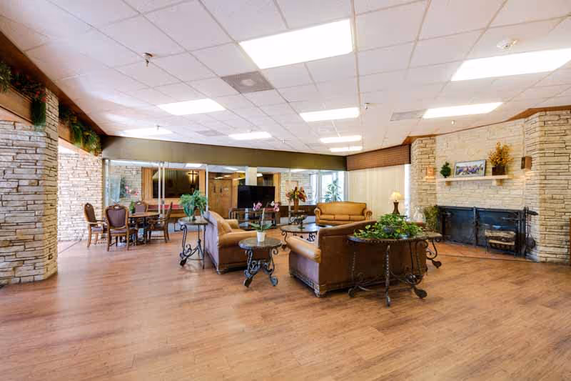 Spacious living room area in a senior living facility with brown leather sofas and armchairs arranged around a coffee table. The room features a stone fireplace with decorative plants and framed pictures on the mantel. There are additional seating areas with wooden chairs and tables near large windows, and the floor is covered with wood-style flooring. The ceiling has recessed lighting panels.