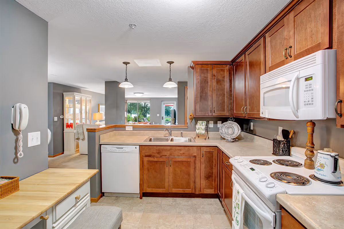 A bright kitchen with wooden cabinets, a white electric stove, microwave, and dishwasher. The kitchen has a beige countertop and a double sink. In the background, there is a view into a living area with large windows showing greenery outside.