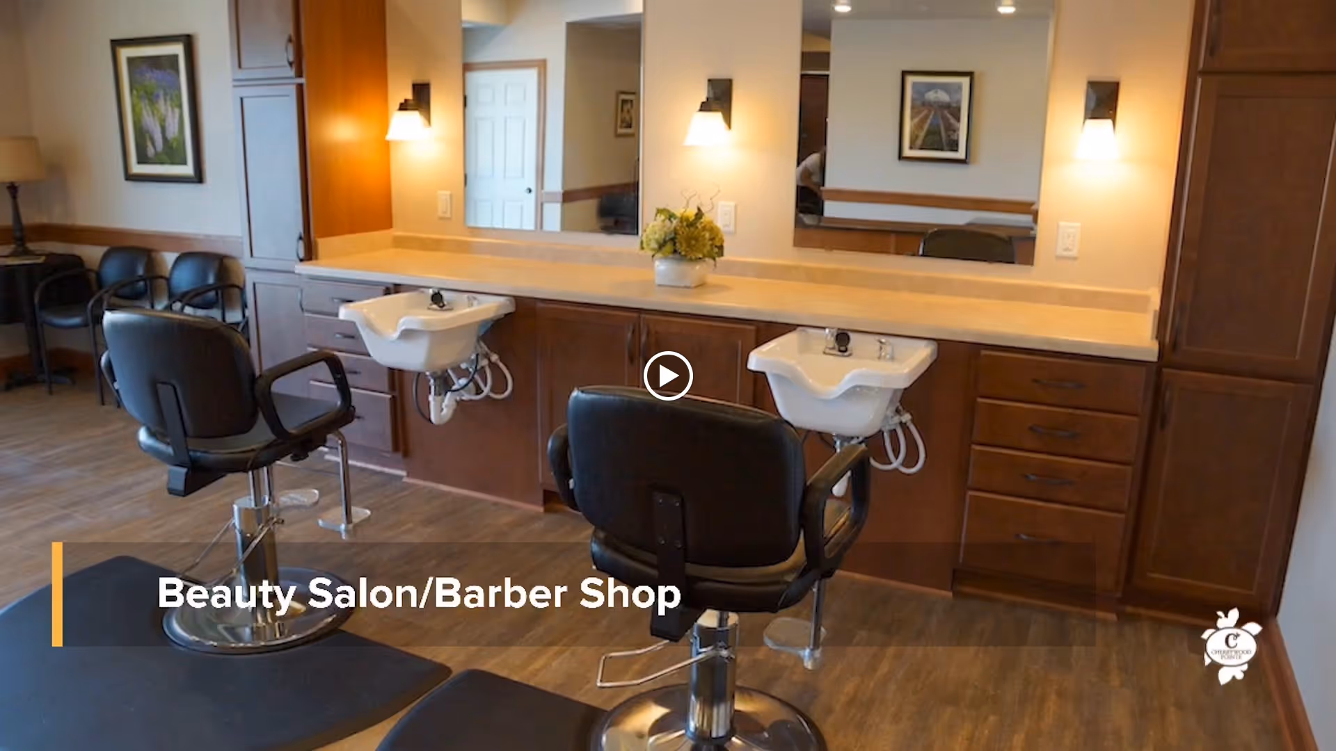 Interior view of a beauty salon/barber shop with two black salon chairs in front of sinks and mirrors. The room has wooden cabinets, a wooden floor, and framed pictures on the walls. There are additional chairs and a lamp in the background.