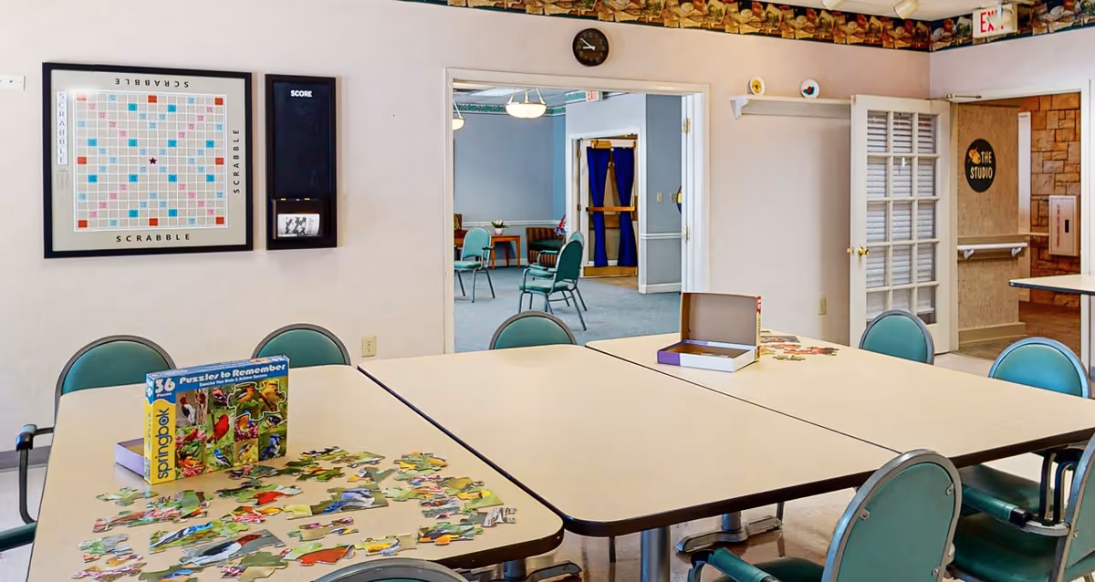A community activity room with tables and green chairs arranged around them. On the tables are puzzle pieces and puzzle boxes. On the wall is a large framed Scrabble board and a score board. Through an open doorway, another room with more chairs and tables is visible. There is a clock above the doorway and a sign on the wall that reads 'The Studio'.