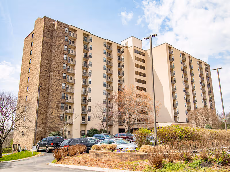 Exterior view of a multi-story senior living facility building with balconies, surrounded by trees and parked cars under a partly cloudy sky.